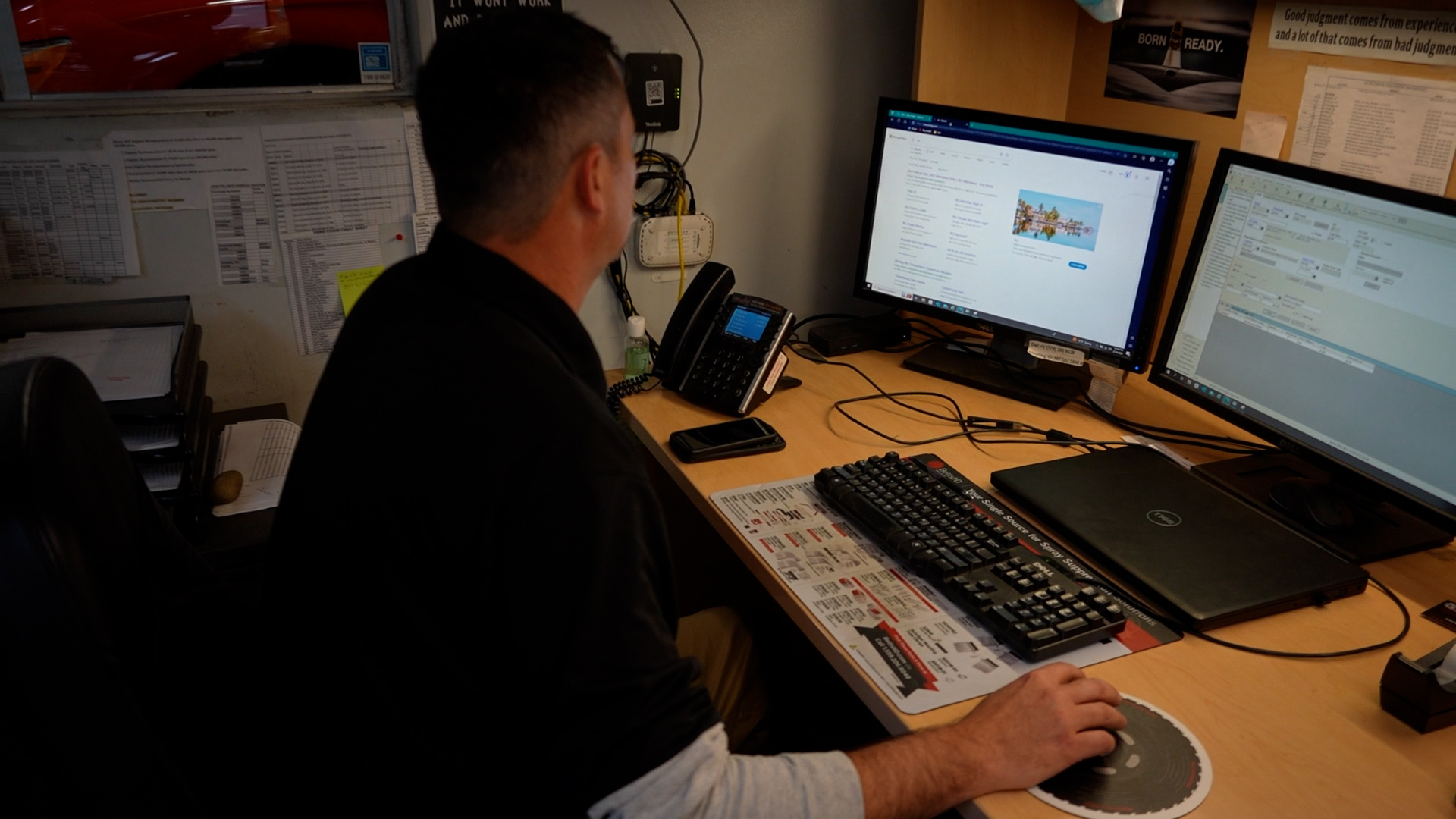 A person working at a desk with dual computer monitors, keyboard, and office equipment in a dimly lit workspace