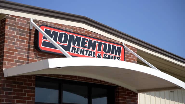 Exterior view of a brick building with a Momentum Rental & Sales sign mounted under a curved awning on a clear blue sky backdrop.